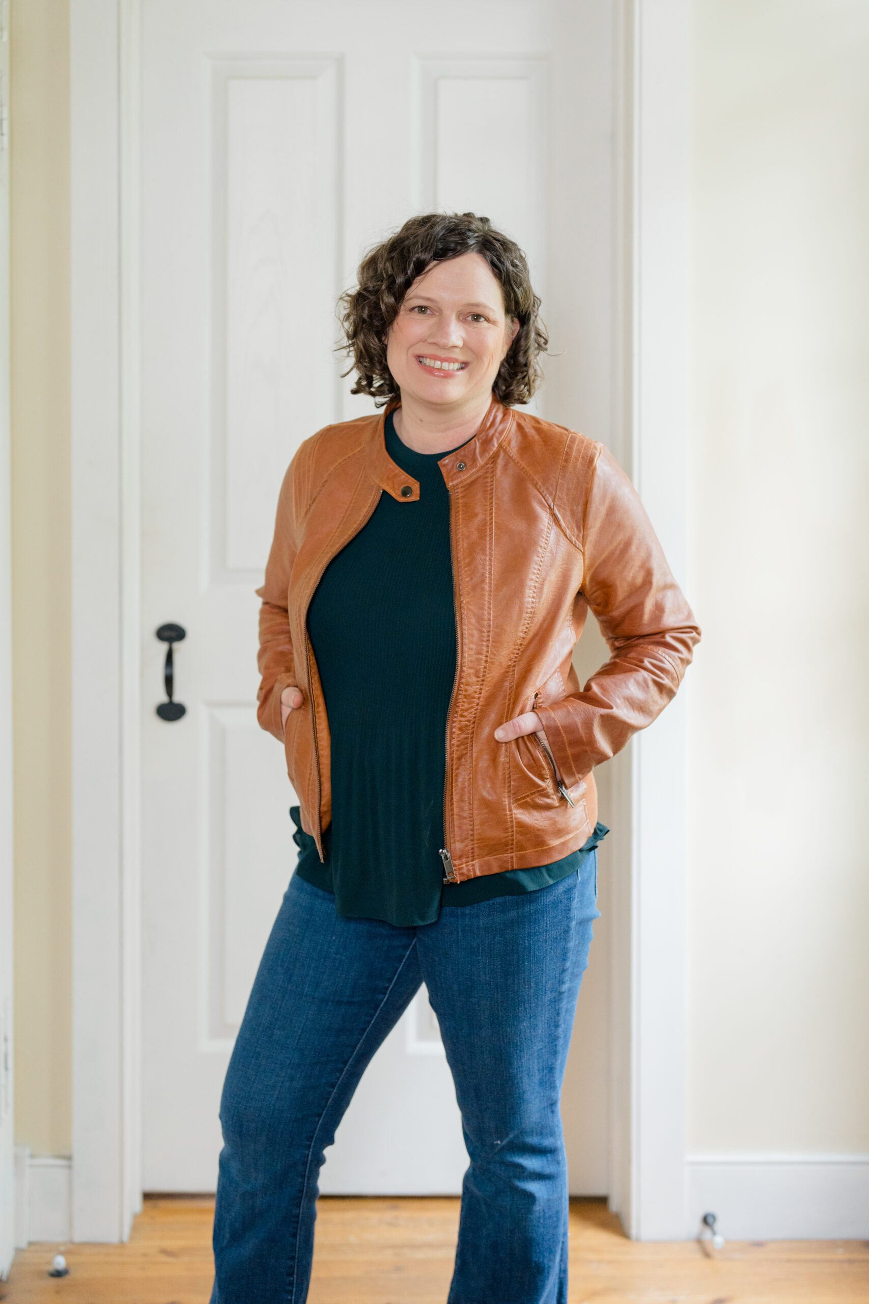 headshot photos with small business owner in dark jeans, a black shirt and a leather jacket standing in a white hallway