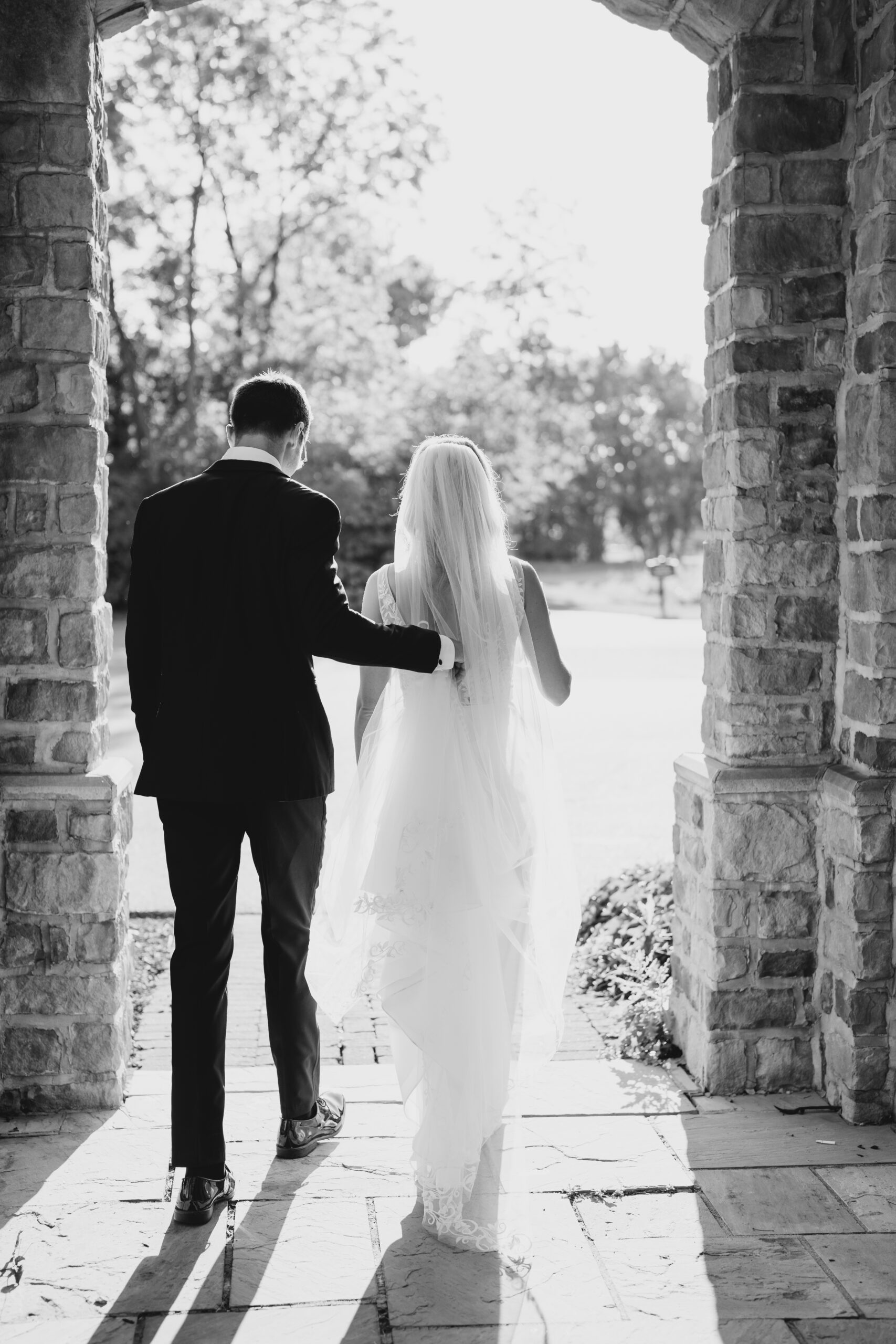 black and white wedding photo of bride and groom walking through stone architecture while the groom holds the brides train and their shadows cast behind them