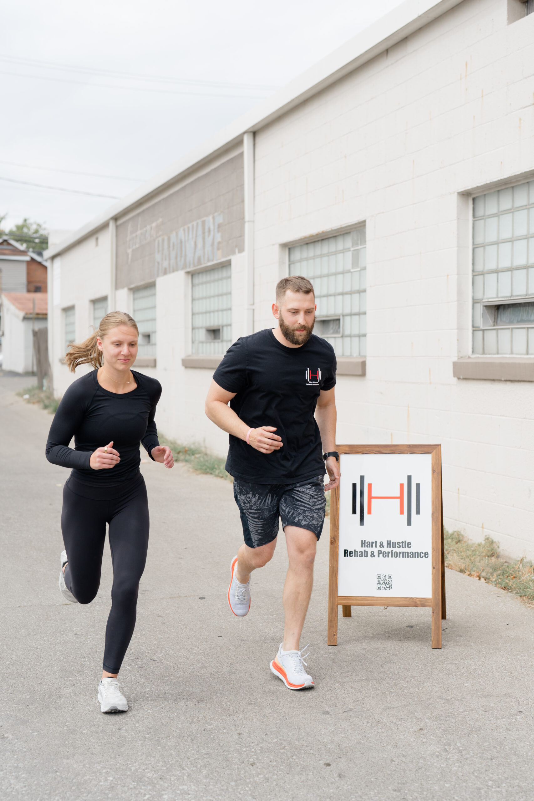 A personal trainer and client run together outside Hart & Hustle Rehab & Performance in Columbus, Ohio. The gym's sign is displayed in the foreground, while the industrial-style building with large glass block windows serves as the backdrop captured by brand photographer