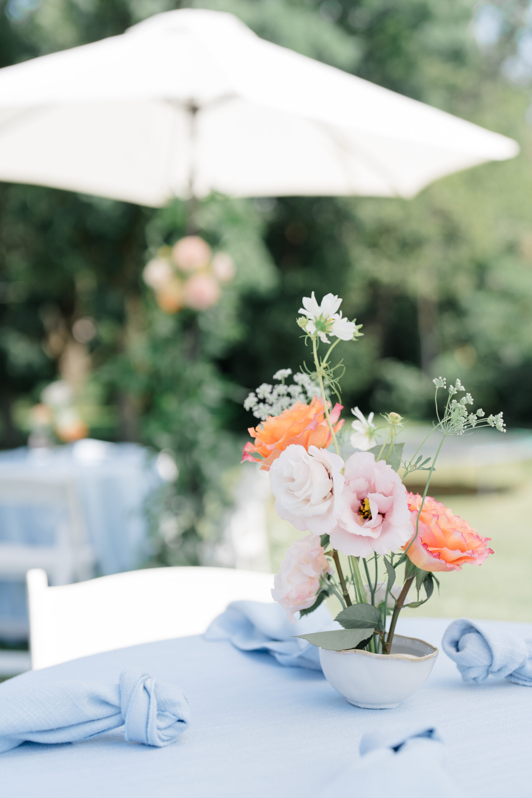 Delicate floral centerpiece with pink, white, and orange blooms in a small ceramic vase, set on a light blue linen table with knotted napkins, ideal for soft, elegant brand photoshoots—photographed by Columbus brand photographer.