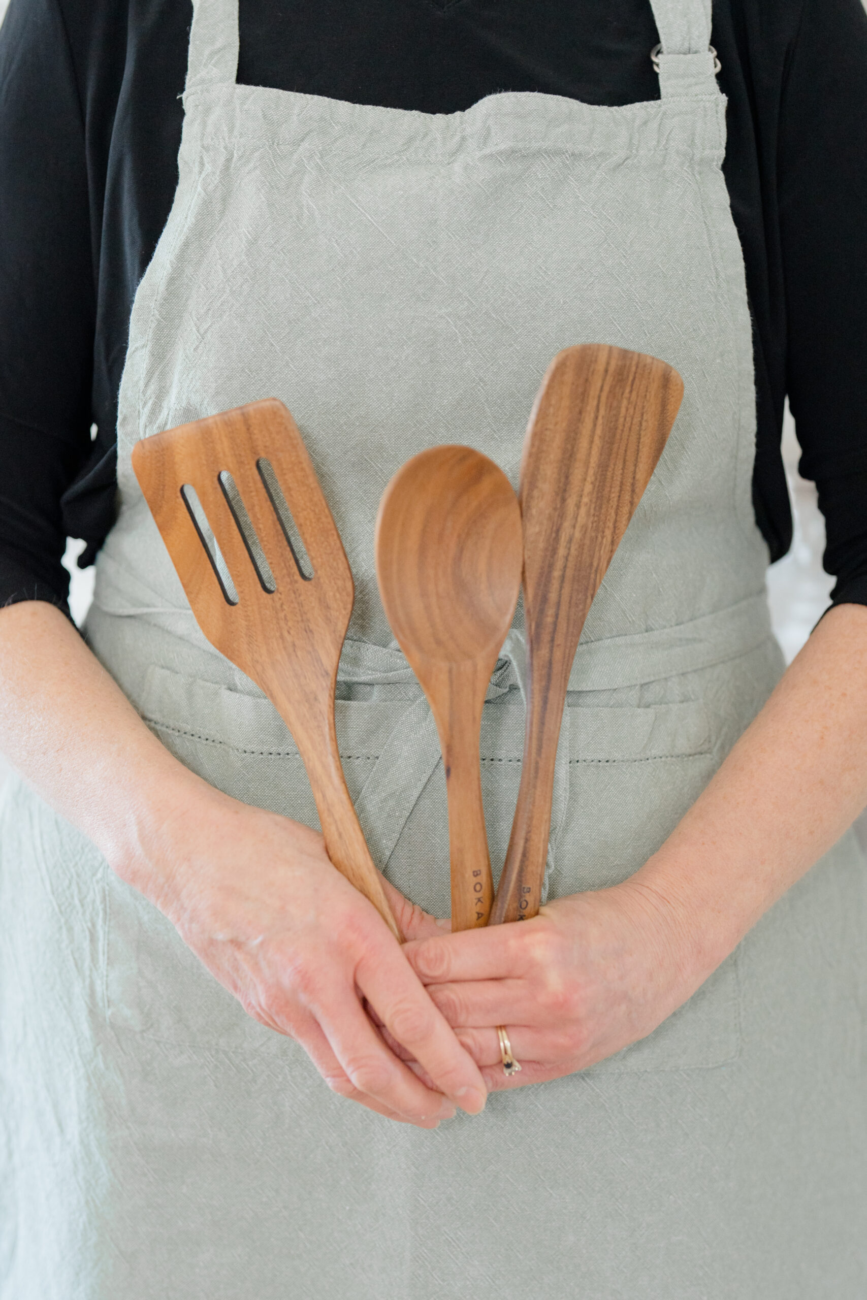 Close-up of a woman in a light gray apron and black shirt holding three wooden kitchen utensils, highlighting personal brand elements for branding in photography.