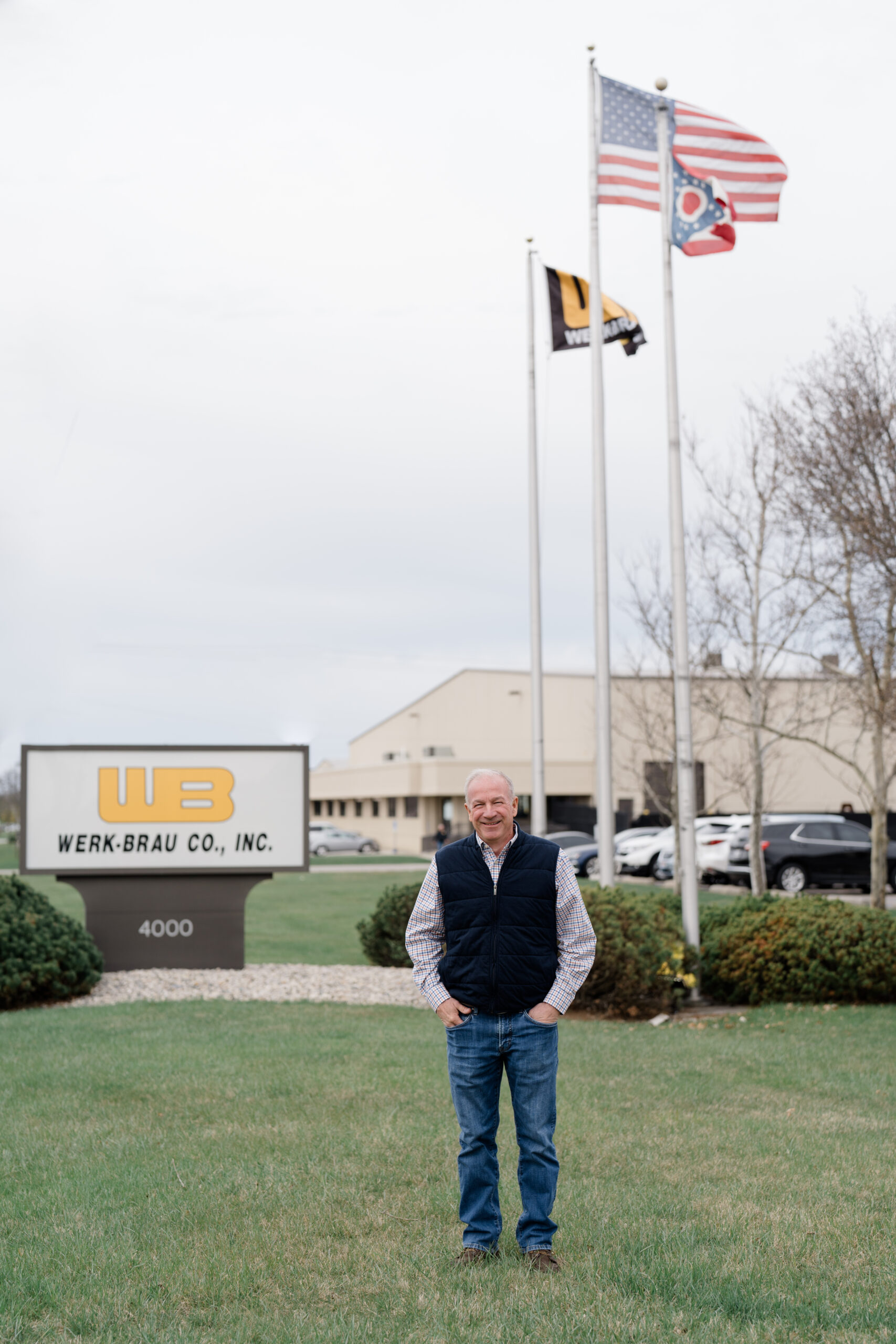 A man standing in front of the Werk-Brau Co., Inc. headquarters with the company sign and three flags—American, Ohio state, and company—visible in the background, reinforcing the brand message of heritage, leadership, and local pride, captured by Columbus brand photographer.